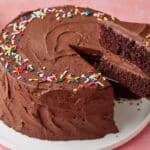 Chocolate Mayonnaise Cake decorated with sprinkles on top. Oe slice being cut, showing the moist crumb and layers of rich buttercream frosting.
