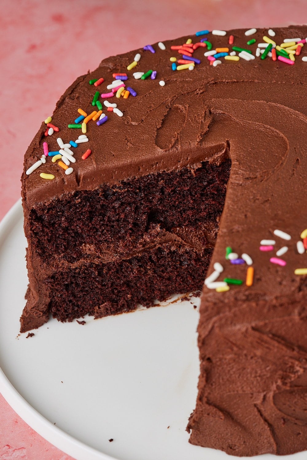Slice removed from a rich Chocolate Mayonnaise Cake frosted with smooth chocolate buttercream and topped with colorful rainbow sprinkles, revealing its moist, tender chocolate crumb and creamy chocolate filling.