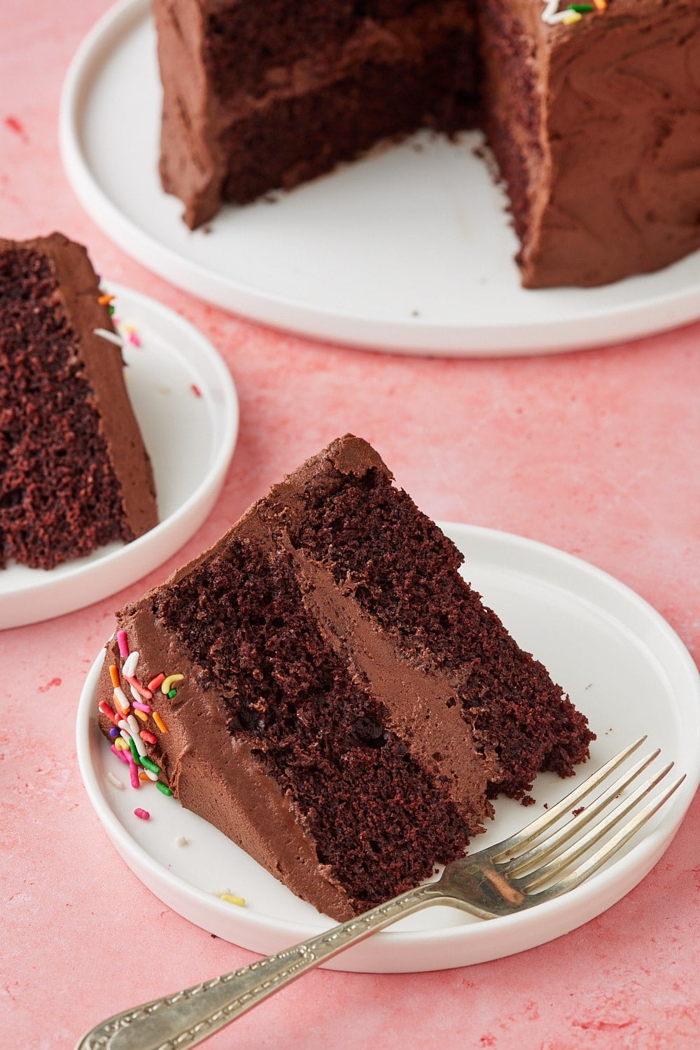 Slice of Chocolate Mayonnaise Cake on a white dessert plate with a fork, showing moist chocolate layers filled and frosted with creamy chocolate buttercream, with the remaining cake in the background.