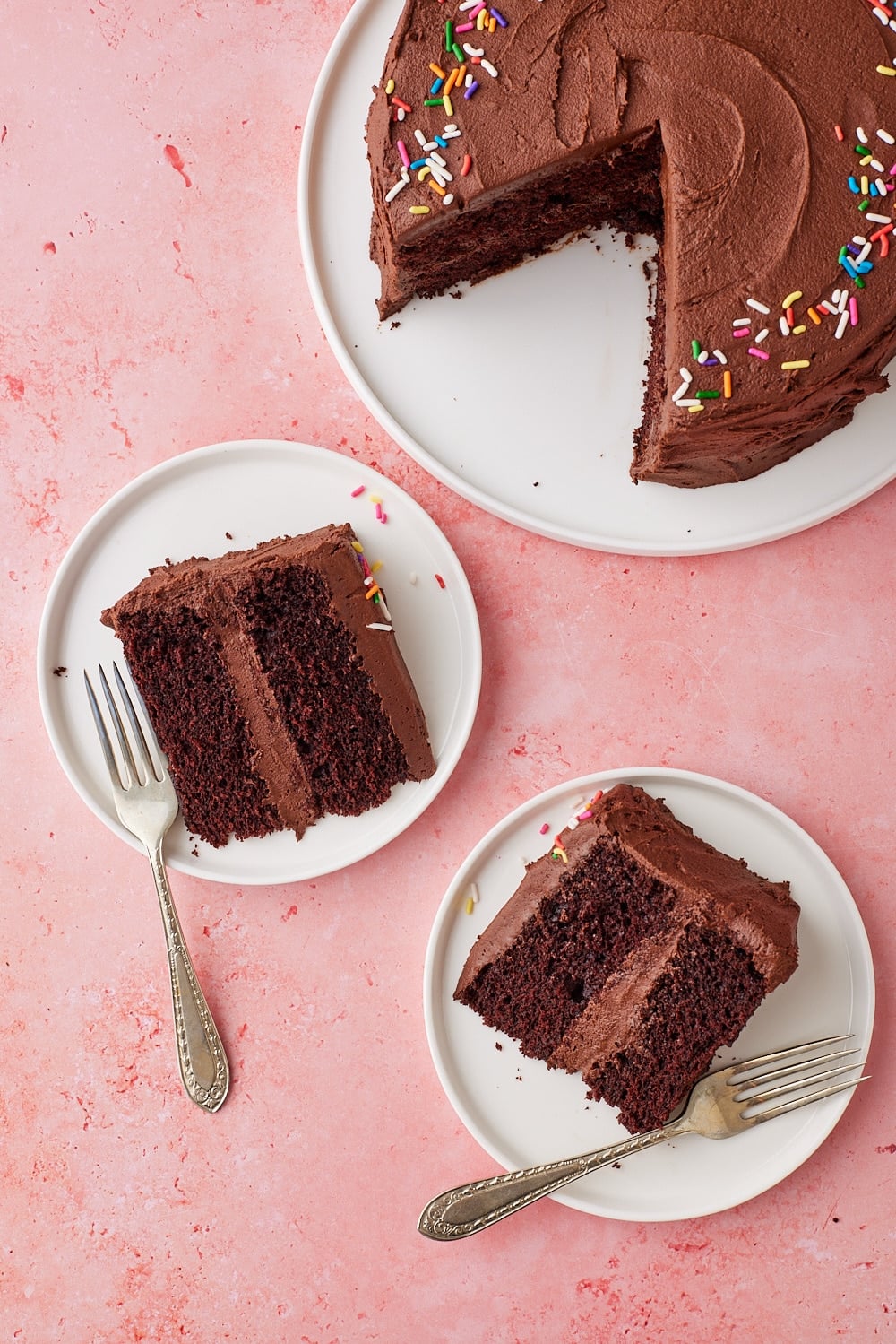 Overhead view of a Chocolate Mayonnaise Cake frosted with chocolate buttercream and decorated with rainbow sprinkles, with two slices served on white dessert plates alongside forks.