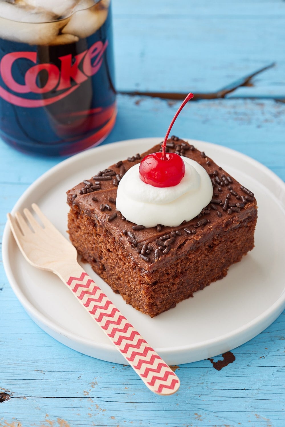 A square of Coca Cola Cake served with whipped cream and cherry on top, with a can of coke on the side. 