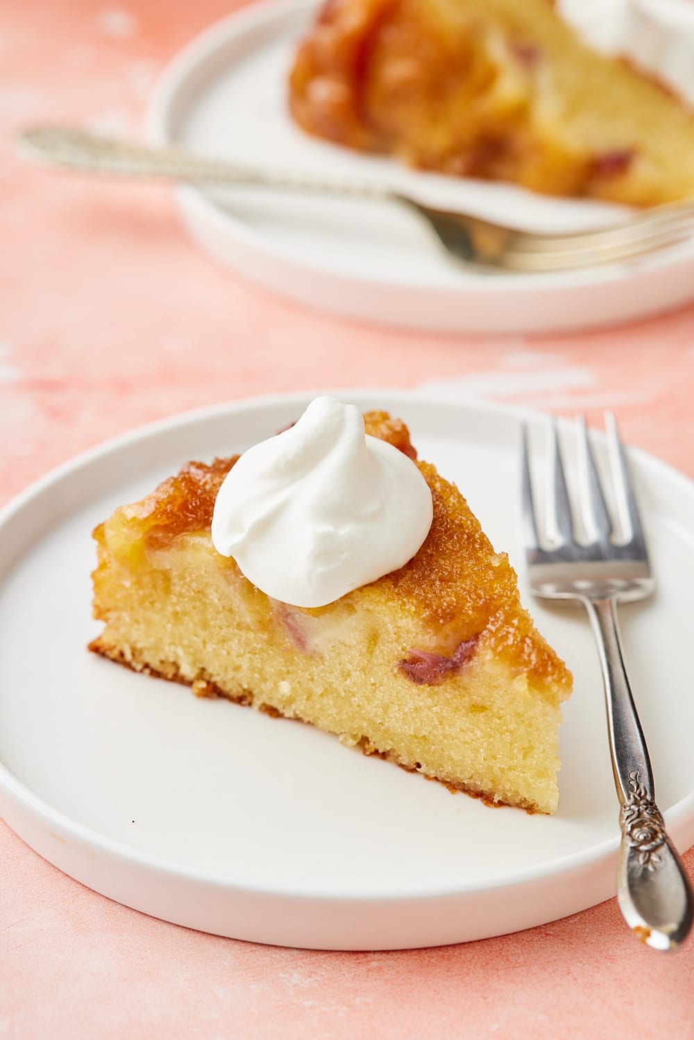 A slice of Peach Upside Down Cake served with softly whipped cream on top. A second slice is in the back. 