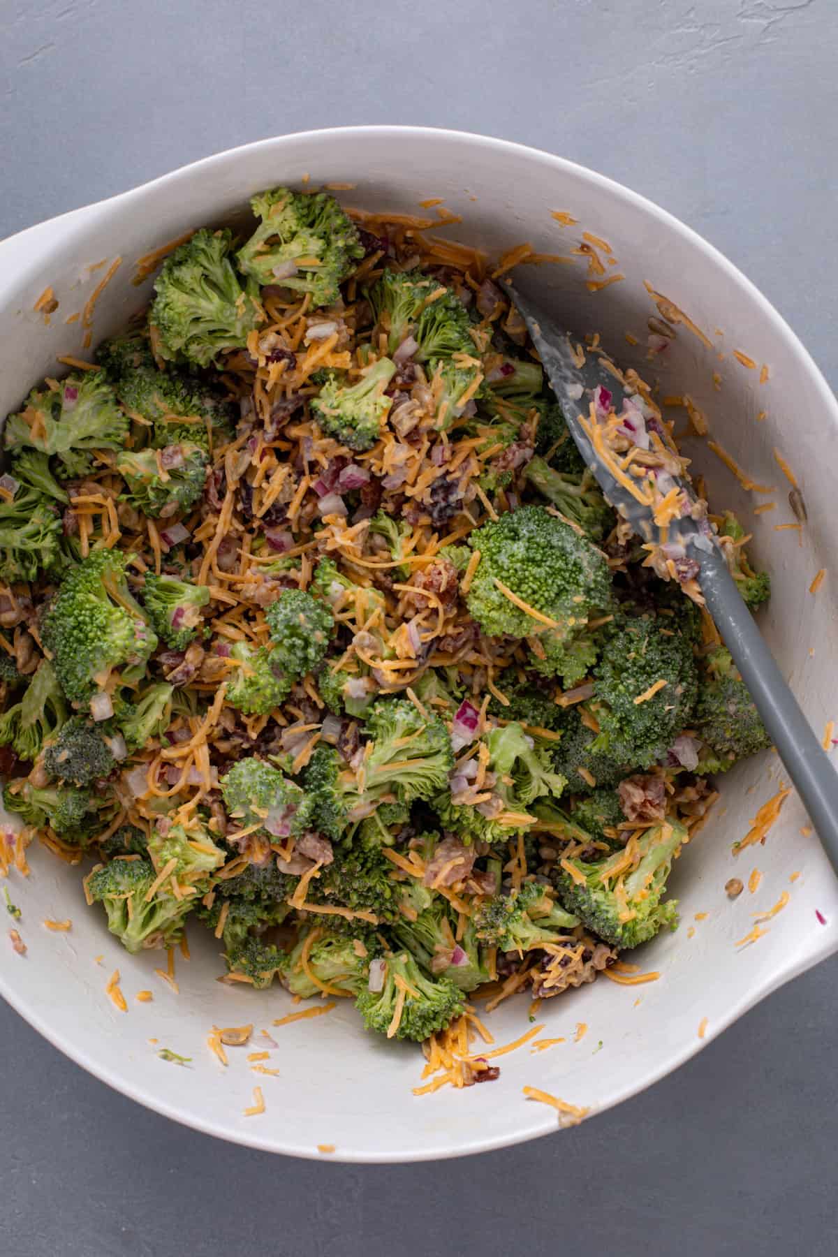 Bacon, cheddar cheese, and sunflower seeds being stirred into broccoli salad.