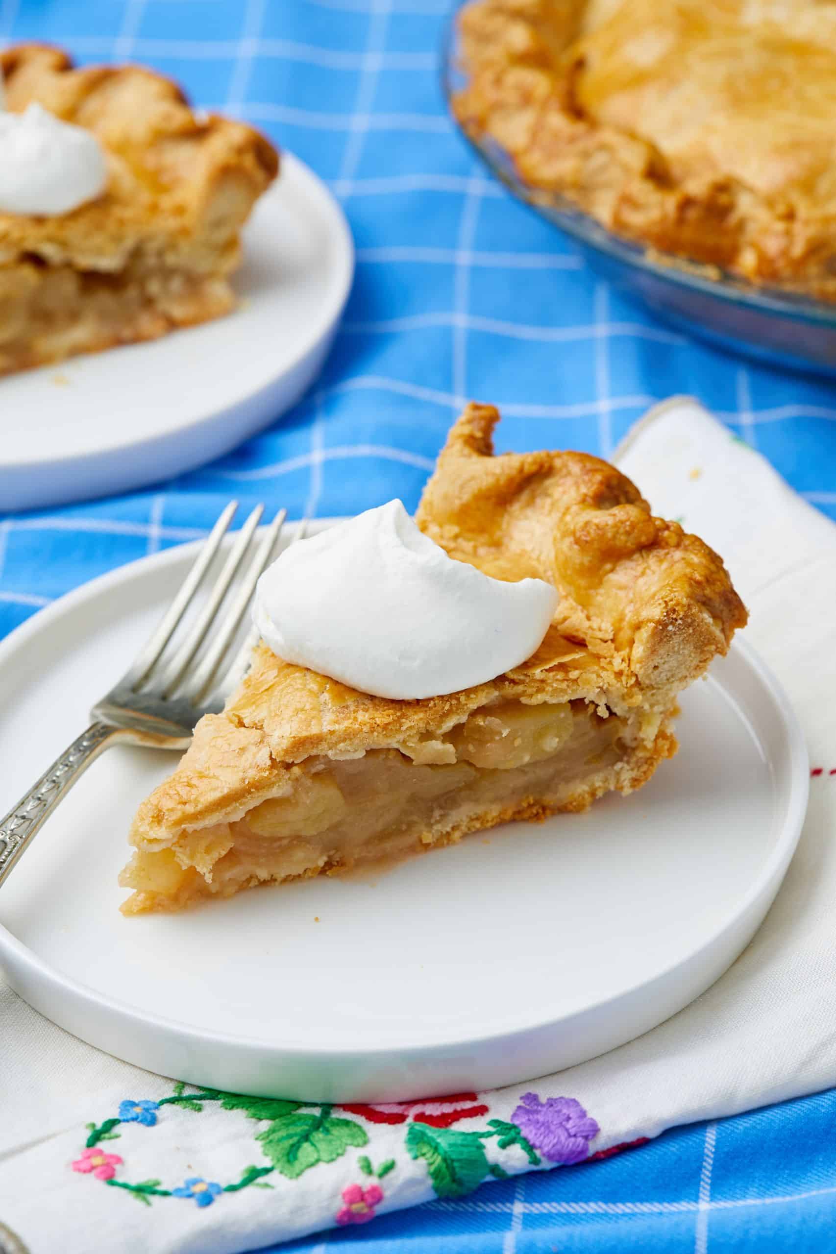 A close up shot at a slice is Traditional Apple Pie, showing the golden flaky crust and soft apples, served with a dollop of whipped cream. 