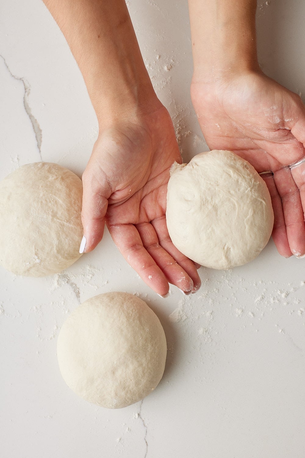 Hands shaping one of four dough balls for Irish batch bread on a lightly floured work surface.